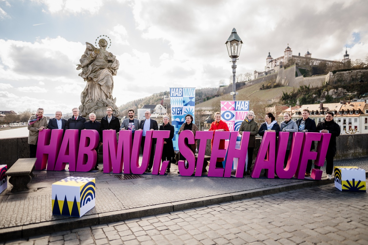 Deutscher Katholikentag - Hab Mut Steh Auf Auf der Mainbrücke stehen Menschen hinter dem Schriftzug "Hab Mut Steh Auf". Im Hintergrund ist die Festung Marienberg zu sehen.