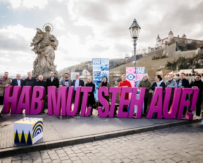 Auf der Mainbrücke stehen Menschen hinter dem Schriftzug "Hab Mut Steh Auf". Im Hintergrund ist die Festung Marienberg zu sehen.